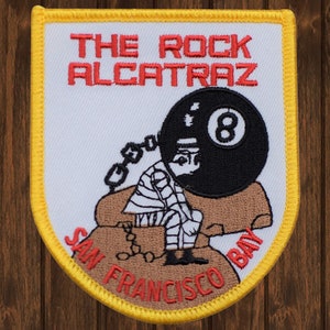 May include: A white embroidered patch with a yellow border. The patch features a cartoon illustration of a prisoner wearing a striped jumpsuit and a ball and chain. The prisoner is sitting on a rock with the number 8 on it. The text "THE ROCK ALCATRAZ" is above the illustration and "SAN FRANCISCO BAY" is below the illustration.