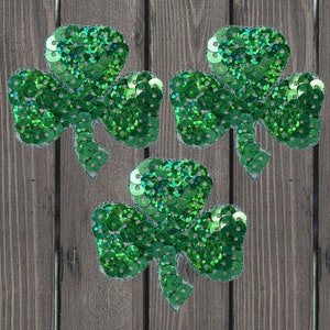 May include: Three green shamrock-shaped patches made of sequins. The patches are on a wood background.