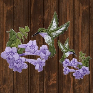 Peut inclure: Deux patchs brodés représentant des colibris et des fleurs violettes. Le plus grand patch montre un colibri en vol avec un corps vert et blanc, tandis que le plus petit patch montre un colibri perché sur une branche avec des feuilles vertes et des fleurs violettes.