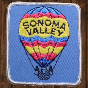 May include: A blue embroidered patch with a hot air balloon. The balloon is yellow, pink, and blue with the words "SONOMA VALLEY" in black.