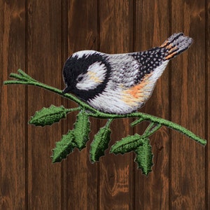 May include: Embroidered patch of a bird on a green branch with leaves. The bird has a black and white head, grey body, and orange details. The patch is designed for application on clothing or accessories.