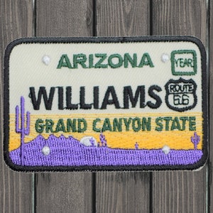 Könnte beinhalten: Ein weißer und grüner gestickter Aufnäher mit dem Text "Arizona" und "Williams, Grand Canyon State". Der Aufnäher zeigt außerdem ein Route 66-Schild und eine Wüstenlandschaft.