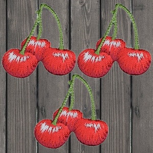 Puede incluir: Tres juegos de cerezas rojas con tallos verdes, cada juego tiene tres cerezas, las cerezas están bordadas sobre un fondo de madera.