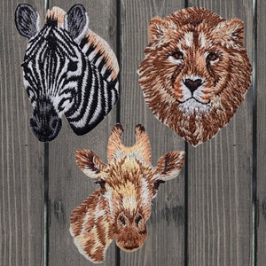 Op de afbeelding: Drie geborduurde patches met dierengezichten: een zebra met zwart-witte strepen, een leeuw met een bruine manen en een giraffe met bruine en beige strepen.