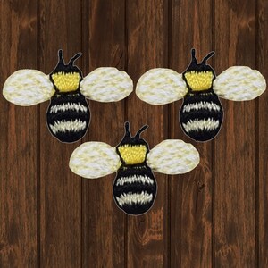 May include: Three embroidered bee patches. Each bee has a yellow head, a black and white striped body, and cream-colored wings. The patches are set against a dark brown wood grain background.