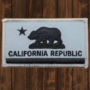 Puede incluir: Un parche bordado blanco y negro con la bandera de la República de California. El parche presenta un oso negro, una estrella y las palabras "CALIFORNIA REPUBLIC".