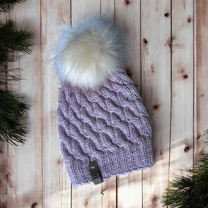 May include: A lavender cable-knit hat with a large, fluffy white and light blue pom-pom. The hat has a ribbed brim and a textured cable pattern. A small tag is attached to the hat. The hat is displayed on a wooden surface.