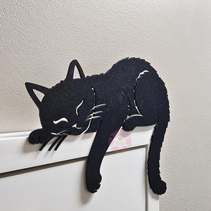 May include: Black cat-shaped wall decor. The cat is lying down with its eyes closed, whiskers, and paws visible. The cat is made of a dark material and is resting on a white surface. The cat's tail hangs down.
