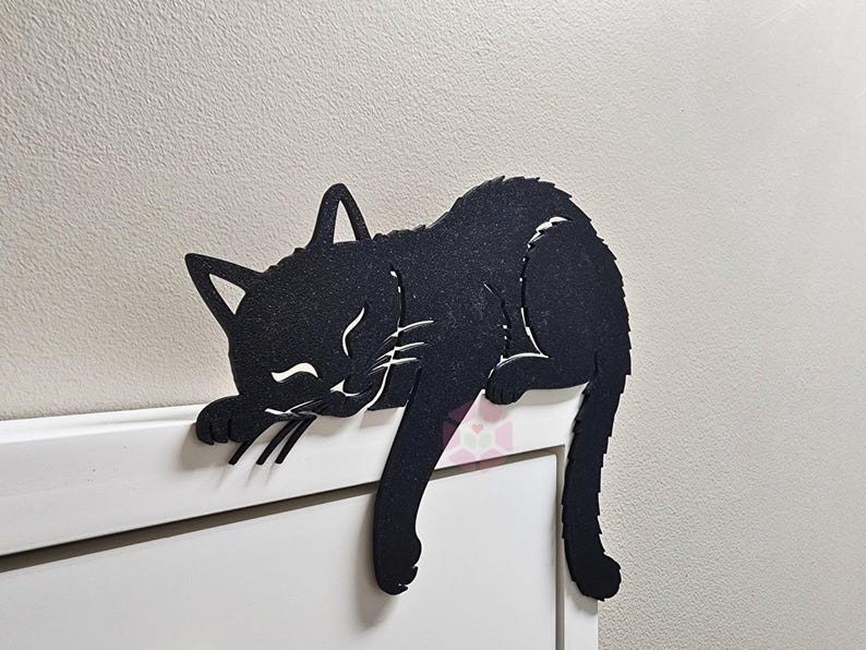 May include: Black cat-shaped wall decor. The cat is lying down with its eyes closed, whiskers, and paws visible. The cat is made of a dark material and is resting on a white surface. The cat's tail hangs down.