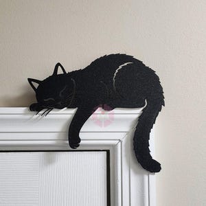 May include: A black cat silhouette decoratively rests on a white door frame. The cat is in a sleeping position, with its head and front paws draped over the edge. The cat's tail curves down, adding to the relaxed pose.