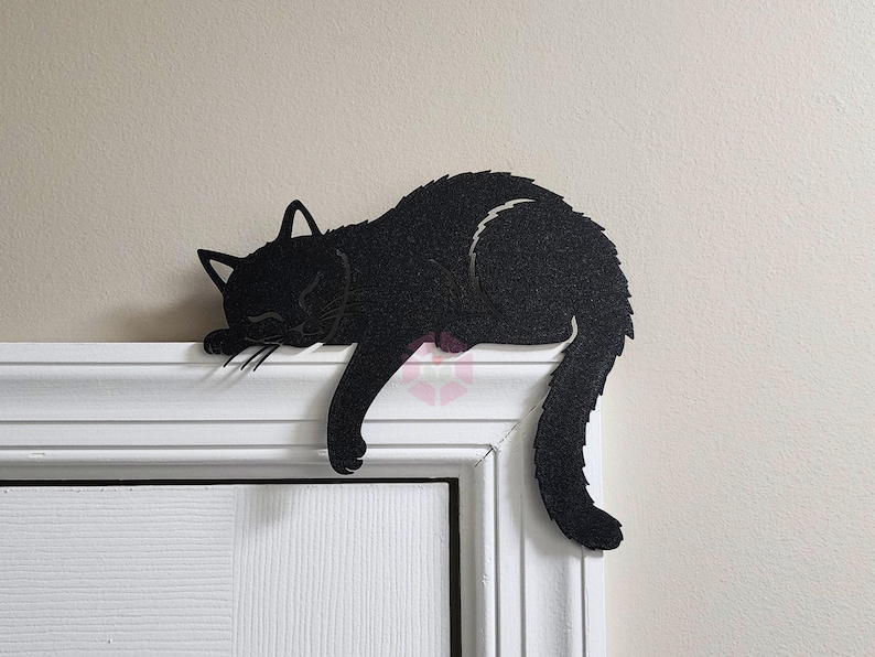 May include: A black cat silhouette decoratively rests on a white door frame. The cat is in a sleeping position, with its head and front paws draped over the edge. The cat's tail curves down, adding to the relaxed pose.