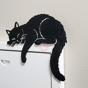 May include: A black cat-shaped decorative item, resting on a white surface. The cat is in a relaxed pose, with its eyes closed and tail curled. The item is made of a solid material, with white accents.