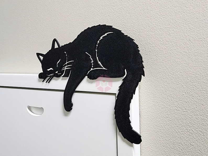 May include: A black cat-shaped decorative item, resting on a white surface. The cat is in a relaxed pose, with its eyes closed and tail curled. The item is made of a solid material, with white accents.