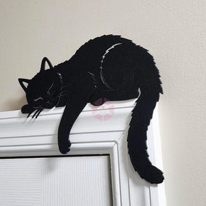 May include: A black cat-shaped decoration resting on a white door frame. The cat is in a sleeping position, with its paws and tail hanging down. The cat's eyes are closed, and its fur is detailed.
