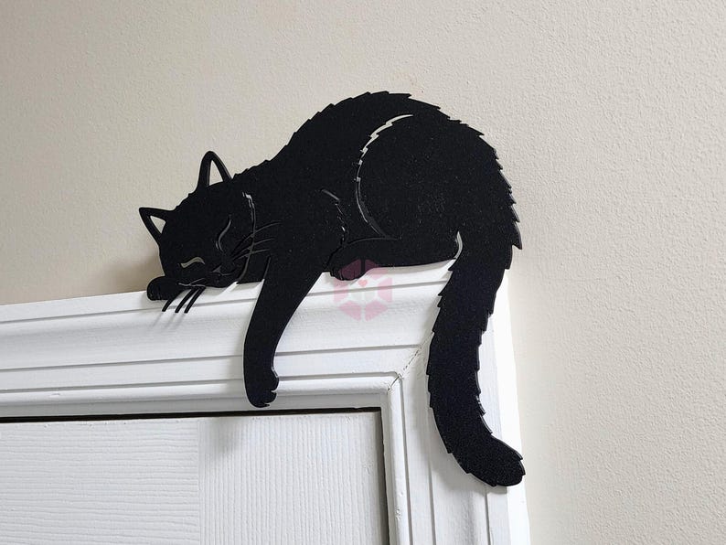 May include: A black cat-shaped decoration resting on a white door frame. The cat is in a sleeping position, with its paws and tail hanging down. The cat's eyes are closed, and its fur is detailed.