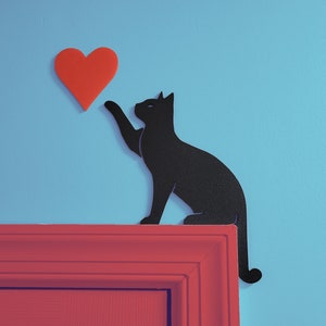May include: A black cat silhouette reaching up to a red heart on a light blue wall. The cat is sitting on a red door frame.