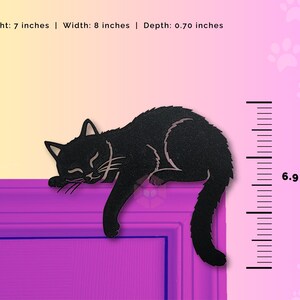 May include: A black cat-shaped decoration, 7 inches high, 8 inches wide, and 0.70 inches deep, is shown resting on a purple surface. The cat is depicted with its eyes closed, suggesting sleep. The background has a gradient of yellow to pink.