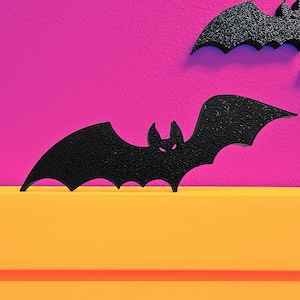 May include: Black bat decorations with textured surfaces are displayed against a vibrant pink background and a yellow surface. The bats have a Halloween theme. The bats are cut out and have a textured surface.