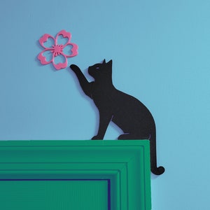 May include: A black silhouette of a cat reaching up to a pink flower on a light blue wall. The cat is sitting on a green trim.