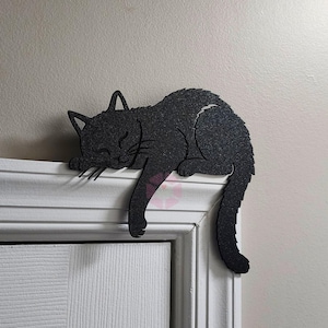 May include: A black cat-shaped decoration is resting on a white door frame. The cat is in a sleeping position, with its head and front paws resting on the frame. The cat's tail hangs down. The cat has a textured surface.