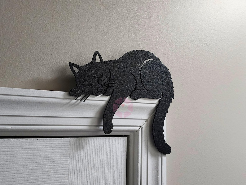 May include: A black cat-shaped decoration is resting on a white door frame. The cat is in a sleeping position, with its head and front paws resting on the frame. The cat's tail hangs down. The cat has a textured surface.