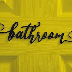 May include: A black metal sign with the word "bathroom" written in a cursive font on a bright yellow wooden door.