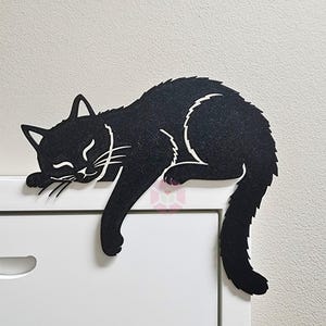 May include: A black cat-shaped felt decoration, lying on a white surface. The cat is in a relaxed pose with its eyes closed and whiskers visible. The cat's tail hangs down, adding a playful touch to the design.