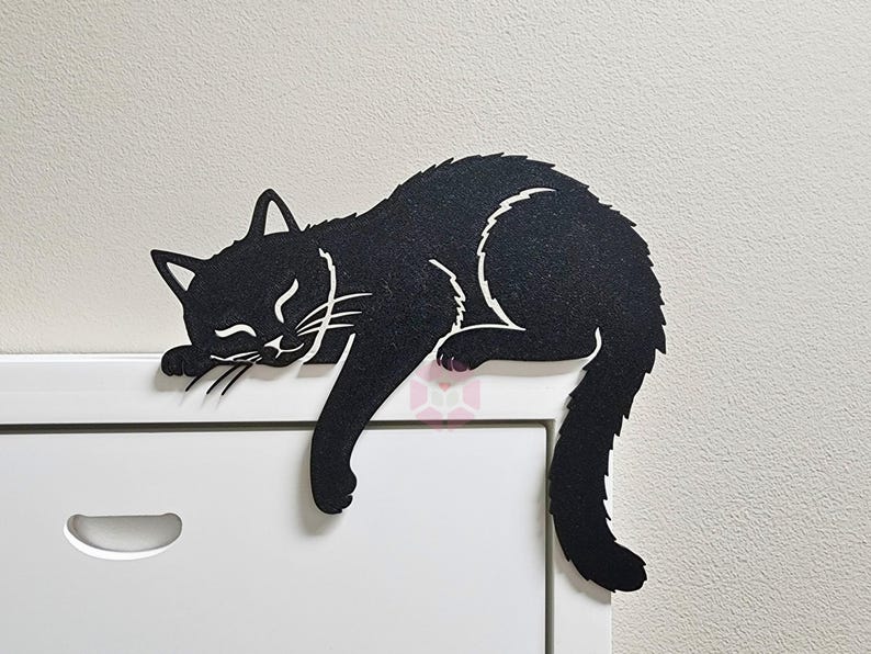 May include: A black cat-shaped felt decoration, lying on a white surface. The cat is in a relaxed pose with its eyes closed and whiskers visible. The cat's tail hangs down, adding a playful touch to the design.