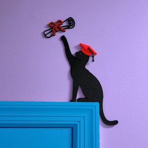 May include: A black cat silhouette holding a red graduation cap and a scroll, mounted on a blue door frame against a purple wall.