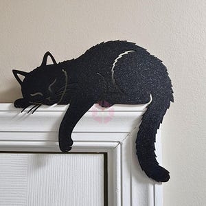 May include: Black metal cat silhouette decor, sleeping on a white door frame. The cat has a textured surface and a curved tail. The cat's eyes are closed, and its whiskers are visible.
