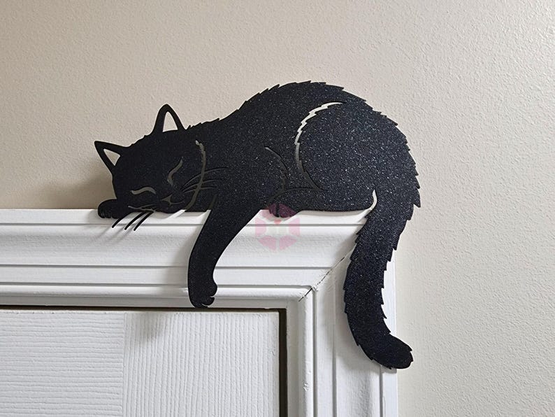May include: Black metal cat silhouette decor, sleeping on a white door frame. The cat has a textured surface and a curved tail. The cat's eyes are closed, and its whiskers are visible.