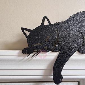 May include: A black metal cat silhouette, sleeping on a white ledge. The cat's body is textured with a glittery finish, and its eyes are closed. The cat's tail hangs down, and its whiskers are visible.