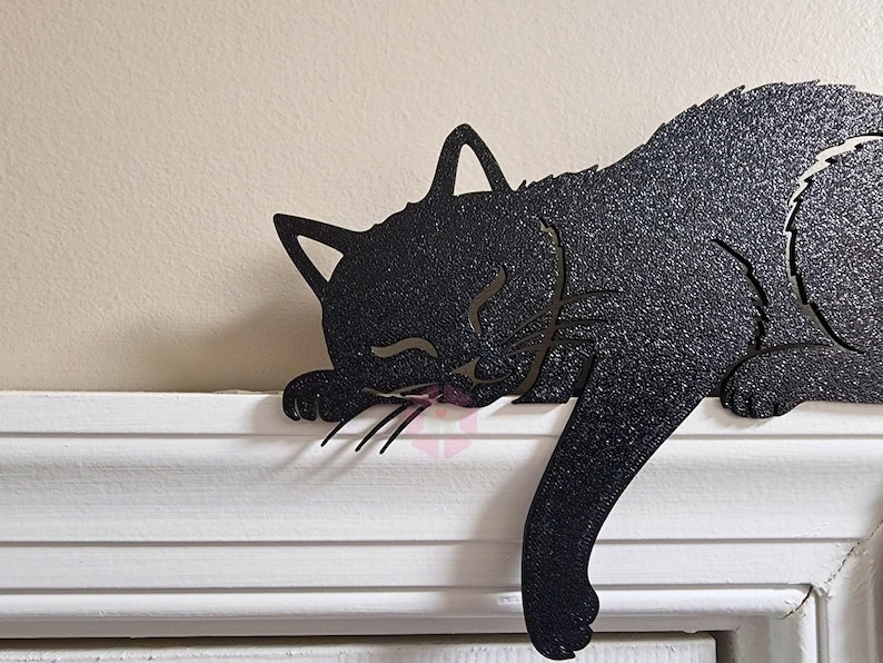 May include: A black metal cat silhouette, sleeping on a white ledge. The cat's body is textured with a glittery finish, and its eyes are closed. The cat's tail hangs down, and its whiskers are visible.