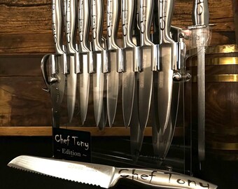 May include: A set of stainless steel kitchen knives, including a bread knife, displayed in a clear acrylic block. The knives have curved handles with the text "Chef Tony" etched on them. The set includes a sharpening steel.