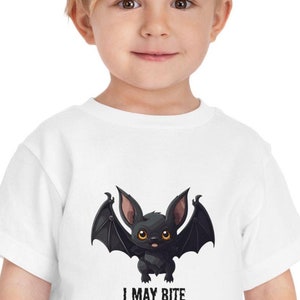 以下が含まれることがあります： 白い子供用Tシャツ。黒いコウモリの漫画のイラストが描かれています。コウモリは黄色い目と広げた翼を持っています。「I MAY BITE」の文字が下にプリントされています。Tシャツは幼い子供が着ています。