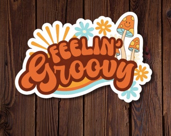 Può includere: Un adesivo con un design retrò che presenta il testo "Feelin' Groovy" in lettere arancioni e marroni su uno sfondo ondulato arancione e giallo. L'adesivo presenta anche funghi e fiori in arancione, giallo e blu.