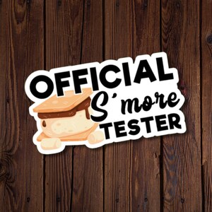May include: A sticker featuring the text "OFFICIAL S'more TESTER" with a cartoon s'more graphic. The s'more includes graham crackers, marshmallow, and chocolate. The background is a dark wood grain texture.