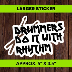 Könnte beinhalten: Ein rechteckiger Aufkleber mit dem schwarzen Text DRUMMERS DO IT WITH RHYTHM in einer fetten, kantigen Schriftart. Hinter dem Text befinden sich zwei braune, kreuzförmig angeordnete Trommelstöcke. Der Aufkleber misst etwa 12,7 cm mal 8,9 cm.