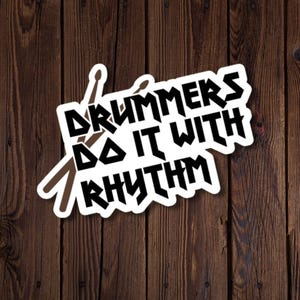 Peut inclure: Un autocollant découpé représentant deux baguettes de batterie marron croisées derrière un texte noir en gras indiquant DRUMMERS DO IT WITH RHYTHM. L'autocollant a une bordure blanche et utilise une police angulaire pour une lecture claire.