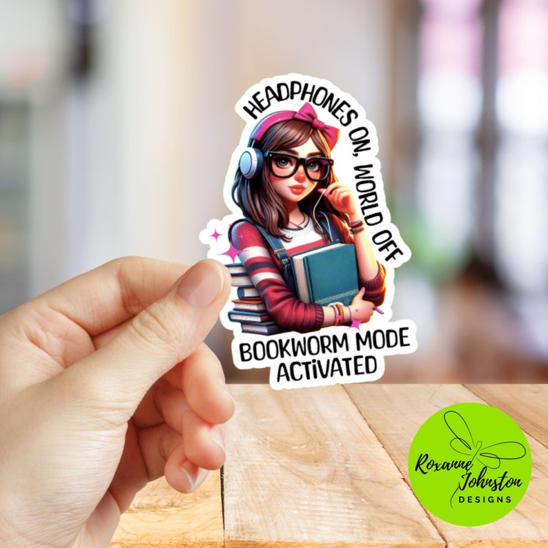 Pu&ograve; includere: Un adesivo con una ragazza dei cartoni animati che indossa delle cuffie e tiene un mucchio di libri. Il testo sull'adesivo recita "Headphones on, World off. Bookworm Mode Activated."