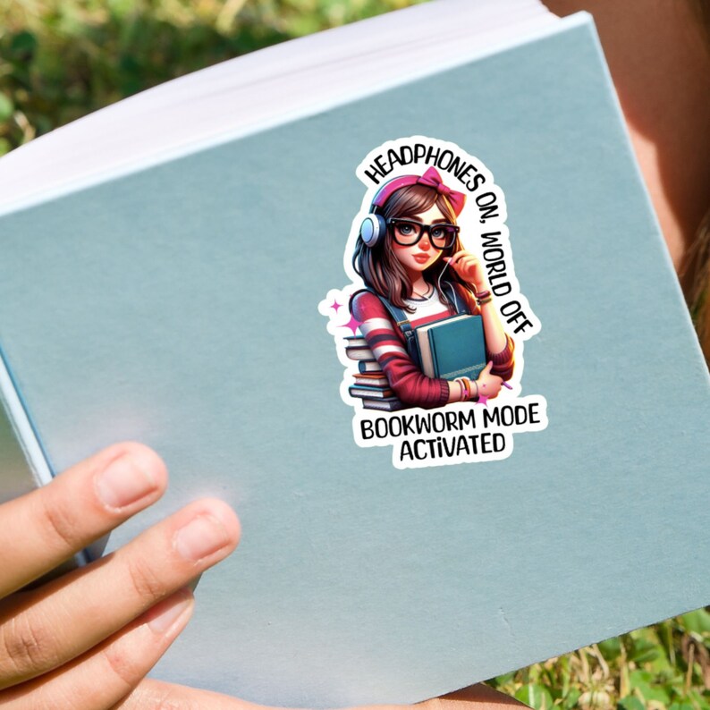 Pu&ograve; includere: Un adesivo con una ragazza dei cartoni animati con le cuffie, che tiene un libro. Il testo dice "Cuffie accese, mondo spento. Modalit&agrave; topo da biblioteca attivata."
