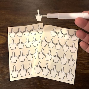 Può includere: Fogli di adesivi con un gesto della mano con il dito medio esteso, in contorno nero su sfondo crema. È presente anche uno stilo bianco. Gli adesivi sono disposti su una superficie marrone scuro.