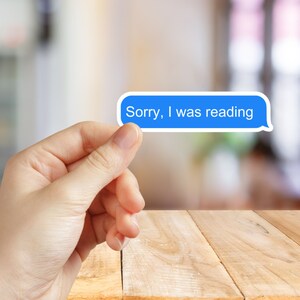 May include: A blue speech bubble sticker with the text "Sorry, I was reading" in white. The sticker is held between fingers against a blurred background and a wooden table.
