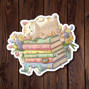 May include: A sticker featuring a cartoon cat napping on a stack of colorful books. The cat is light orange with darker orange stripes. A vase of flowers and two blue birds are also included in the design.