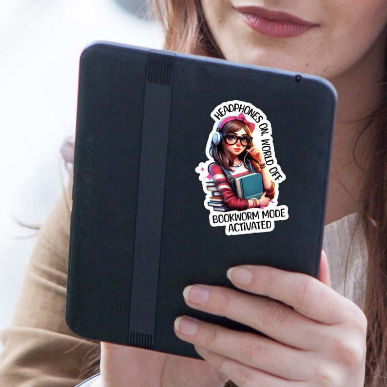 Pu&ograve; includere: Un adesivo con una ragazza dei cartoni animati che indossa le cuffie e tiene un mucchio di libri. Il testo sull'adesivo dice "Headphones on, World off. Bookworm Mode Activated."