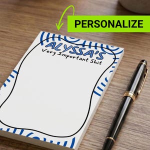 May include: A white notepad with blue decorative accents and the text "ALYSSA'S Very Important Shit". A black pen with gold accents rests on a wooden surface next to the notepad. The word "PERSONALIZE" is in a green box.