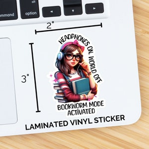 Pu&ograve; includere: Un adesivo in vinile laminato con una ragazza dei cartoni animati con cuffie rosa, occhiali e una pila di libri. Il testo recita "Headphones On, World Off" e "Bookworm Mode Activated".