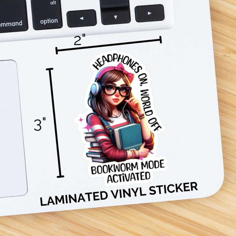 Pu&ograve; includere: Un adesivo in vinile laminato con una ragazza dei cartoni animati con cuffie rosa, occhiali e una pila di libri. Il testo recita "Headphones On, World Off" e "Bookworm Mode Activated".