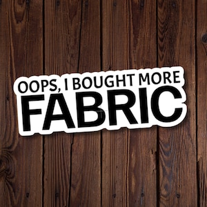 Può includere: Un adesivo bianco con la scritta "OOPS, I BOUGHT MORE FABRIC" in grassetto, nero. L'adesivo è posto su uno sfondo di assi di legno verticali, marrone scuro.