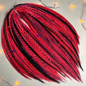 Puede incluir: Un conjunto de rastas rojas, negras y burdeos. Las rastas están adornadas con costuras decorativas y pequeñas cuentas de colores. Las extensiones de cabello están dispuestas en forma de abanico.